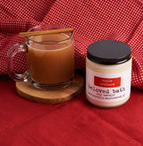 Apple Orchard Candle next to a cup of warm cider with a cinnamon stick. The cider cup is clear and is on a round piece of wood. surrounded by a red and white check cloth and sitting on a red cloth.